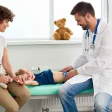 Lekarz bada brzuszek dziecka, które leży na kozetce. Mama z pluszowym misiem obok. Dziecko uśmiecha się, mimo że może mieć biegunkę u niemowlaka.