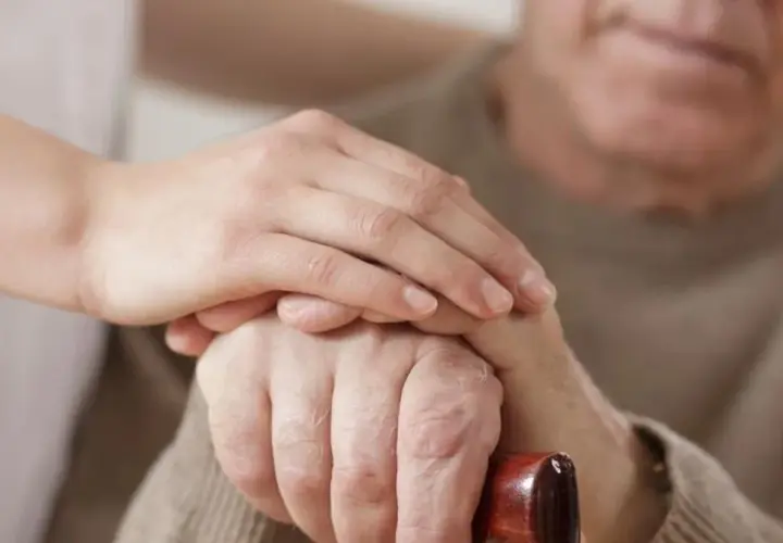 Dłoń opiekunki na dłoni starszego mężczyzny, który trzyma laskę. Wsparcie i troska, jak przy leczeniu chorób, np. parkinsonizmu, gdzie pomocne bywa Madopar.