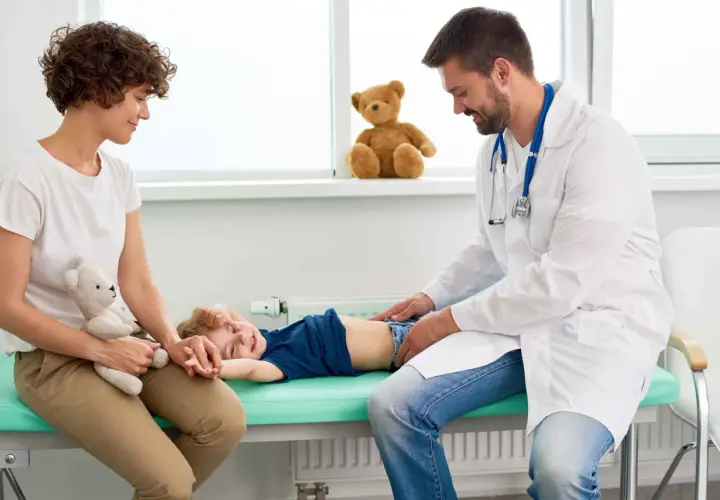 Lekarz bada brzuszek dziecka, które leży na kozetce. Mama z pluszowym misiem obok. Dziecko uśmiecha się, mimo że może mieć biegunkę u niemowlaka.