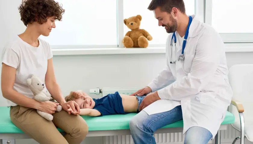 Lekarz bada brzuszek dziecka, które leży na kozetce. Mama z pluszowym misiem obok. Dziecko uśmiecha się, mimo że może mieć biegunkę u niemowlaka.