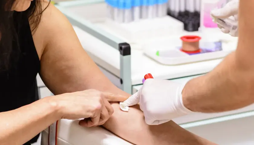 Pobieranie krwi do badań. Ręka pacjentki z widocznymi żyłami, pielęgniarka w białej rękawiczce przygotowuje miejsce wkłucia. W tle probówki i sprzęt medyczny. MPV niskie.